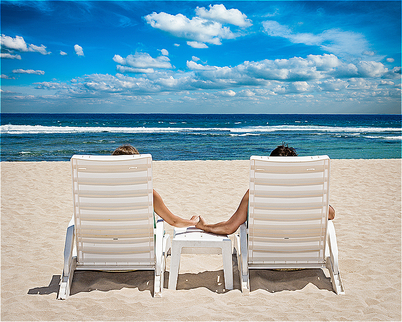rear view of couple sitting in beach chairs on the beach and holding hands while looking at the ocean